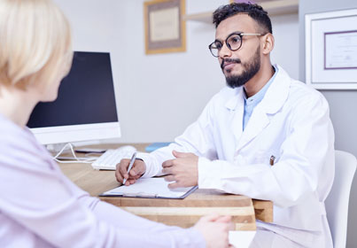 Male dentist in white coat and glasses consulting with female patient at desk in dental office.