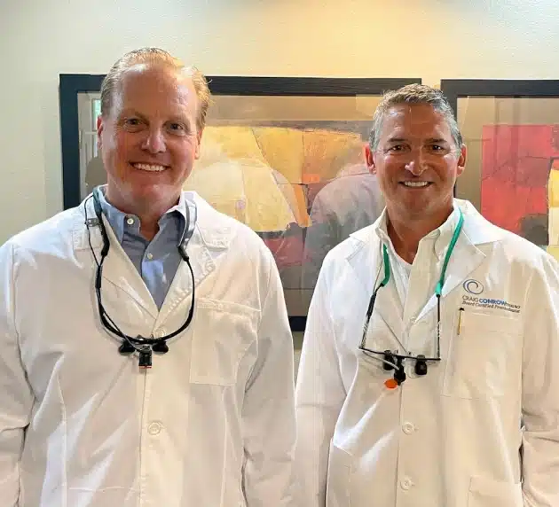 Two male dentists in white coats smiling at camera in dental office setting.