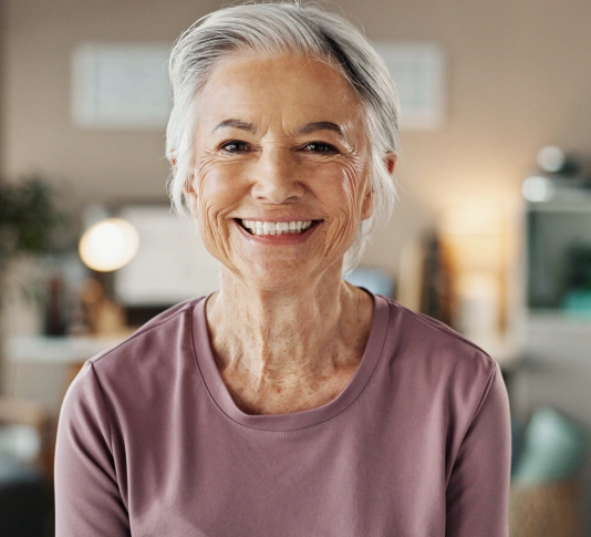 Smiling senior woman in purple shirt, indoor home setting with blurred background.